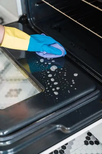 tidying the kitchen by cleaning the oven using rubber gloves and a cloth