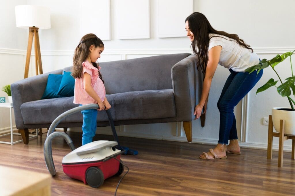 a woman and a young girl working together to vacuum under the couch.